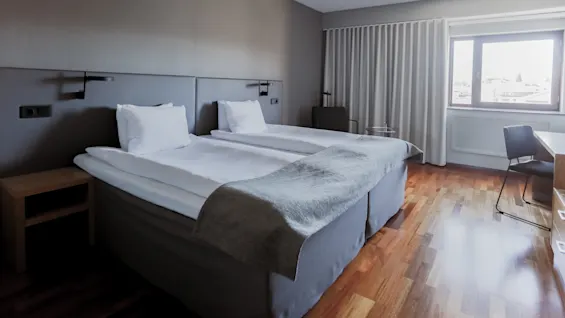 Two neatly made twin beds sit side by side in a minimalistic hotel room with wooden floors, a bedside table, a large window with curtains, and a desk with a chair.
