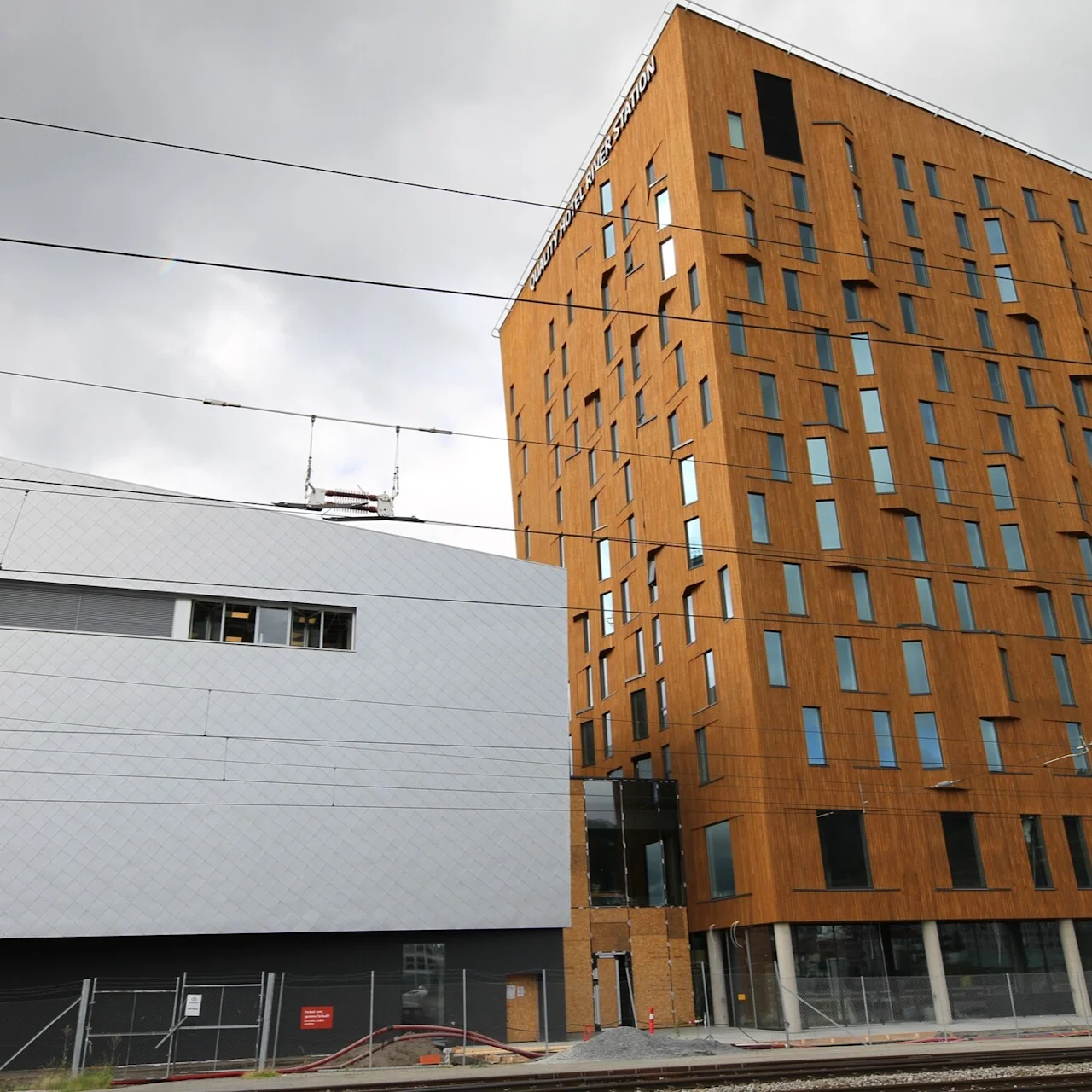 En høy, moderne trekonstruksjon med uregelmessige vinduer står ved siden av en skrånende hvit bygning, omgitt av grå himmel og togskinner. Tekst: "Mjøstårnet".