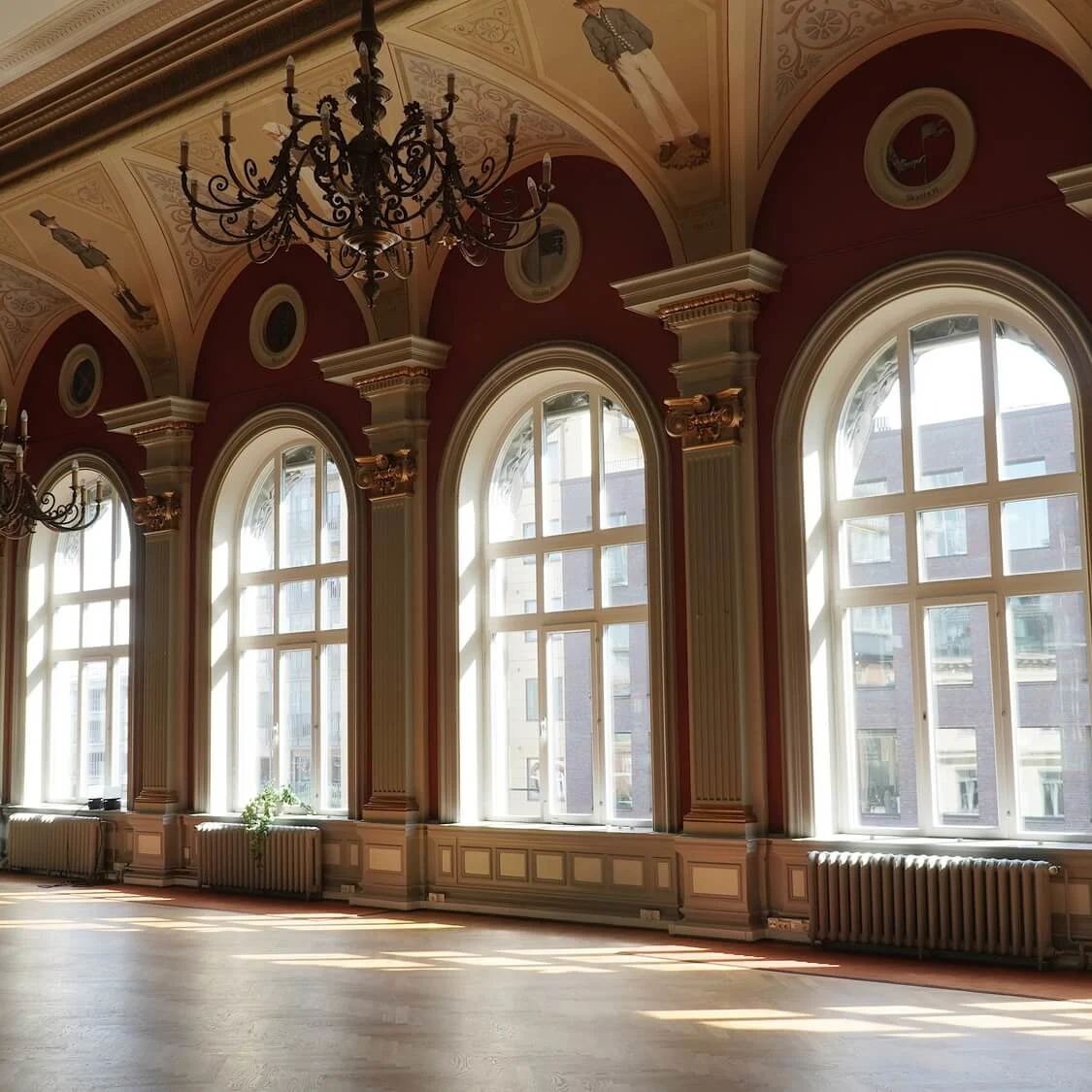 An elegant ballroom with high ceilings, ornate chandeliers, and tall arched windows casting sunlight on polished wooden floors.
