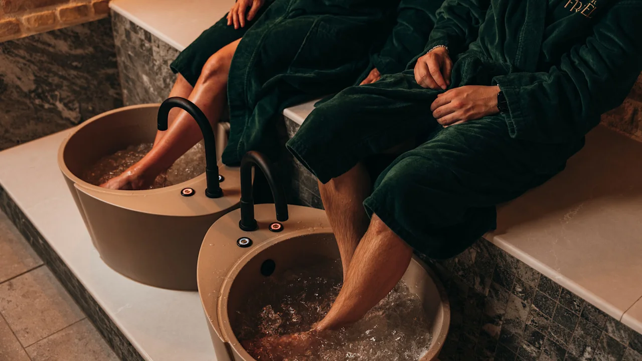 Two people in green robes soak their feet in bubbling foot baths, seated on a tiled bench in a spa setting. "FRIÉL" is embroidered on their robes.