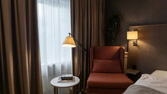 A cozy corner in a hotel room at Home Hotel Grand Bodø with an armchair, reading lamp, and a window with curtains.
