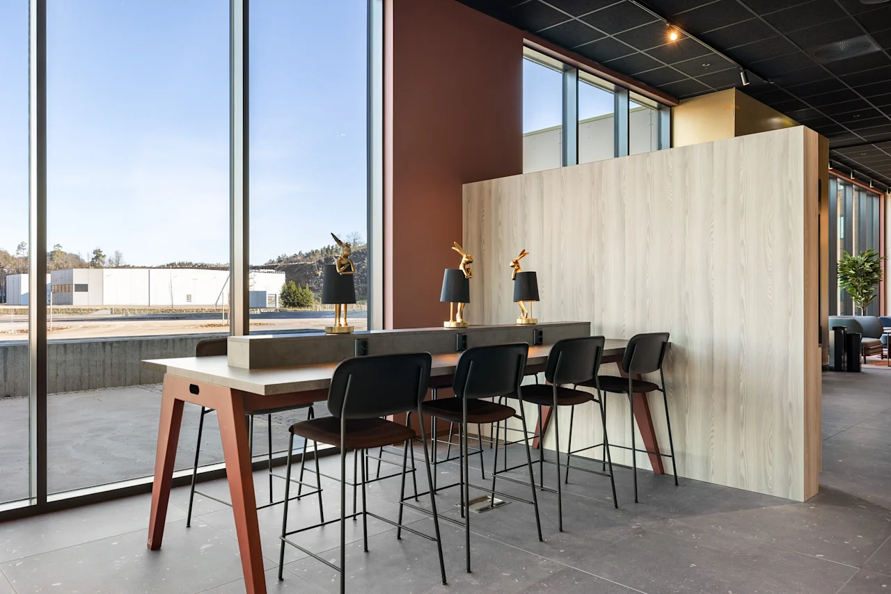 Moderne hotellobby med et langt høyt bord og barkrakker ved store vinduer, som tilbyr en lys arbeidsplass.