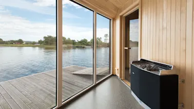 Innendørs sauna med store glassvinduer som vender mot en rolig innsjø med trær i det fjerne. Vegger og gulv i tre skaper en varm, fredfull atmosfære.