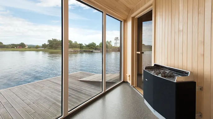 An indoor sauna features large glass windows overlooking a serene lake with trees in the distance. The wooden walls and floor create a warm, peaceful atmosphere.