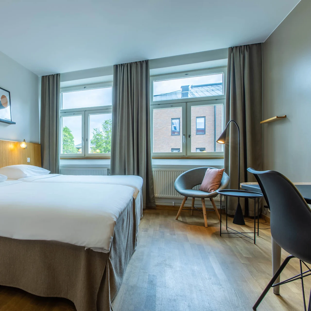 A neatly made bed in a modern hotel room with art on the wall, a sitting area, and a view of a city through the window.