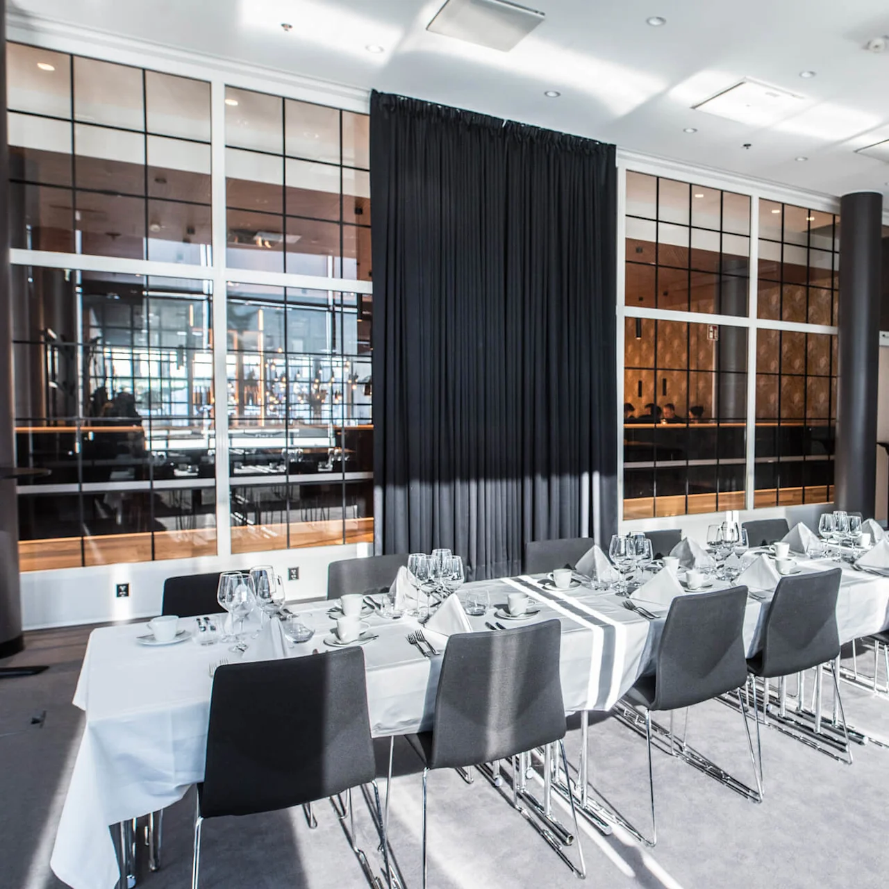 A long, elegantly set dining table with white tablecloth and black chairs stands in a modern, well-lit room with large windows, patterned wallpaper, and a black curtain.