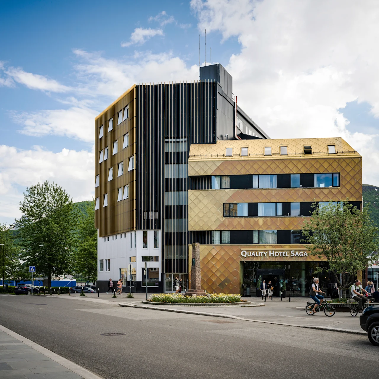 A building labeled "QUALITY HOTEL SAGA" stands in a bustling street context, featuring modern architecture with reflective panels and multiple windows; pedestrians and cyclists move nearby.