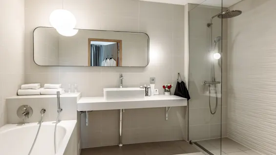 Bathtub with chrome faucet lies beside sink and mirror; towels neatly stacked, and shower enclosed in glass. Soft lighting creates a serene, modern bathroom ambience.
