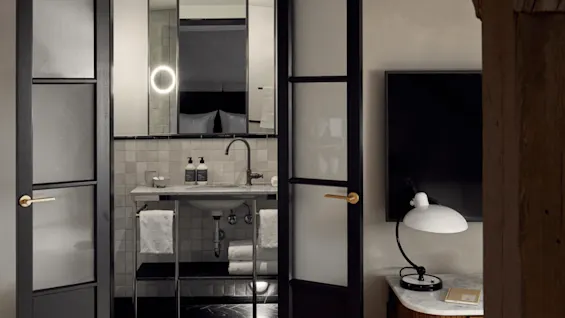 A bathroom with an open doorway reveals a modern sink, two soap dispensers, and a mirror reflecting a halo light, accompanied by folded towels on a lower shelf.