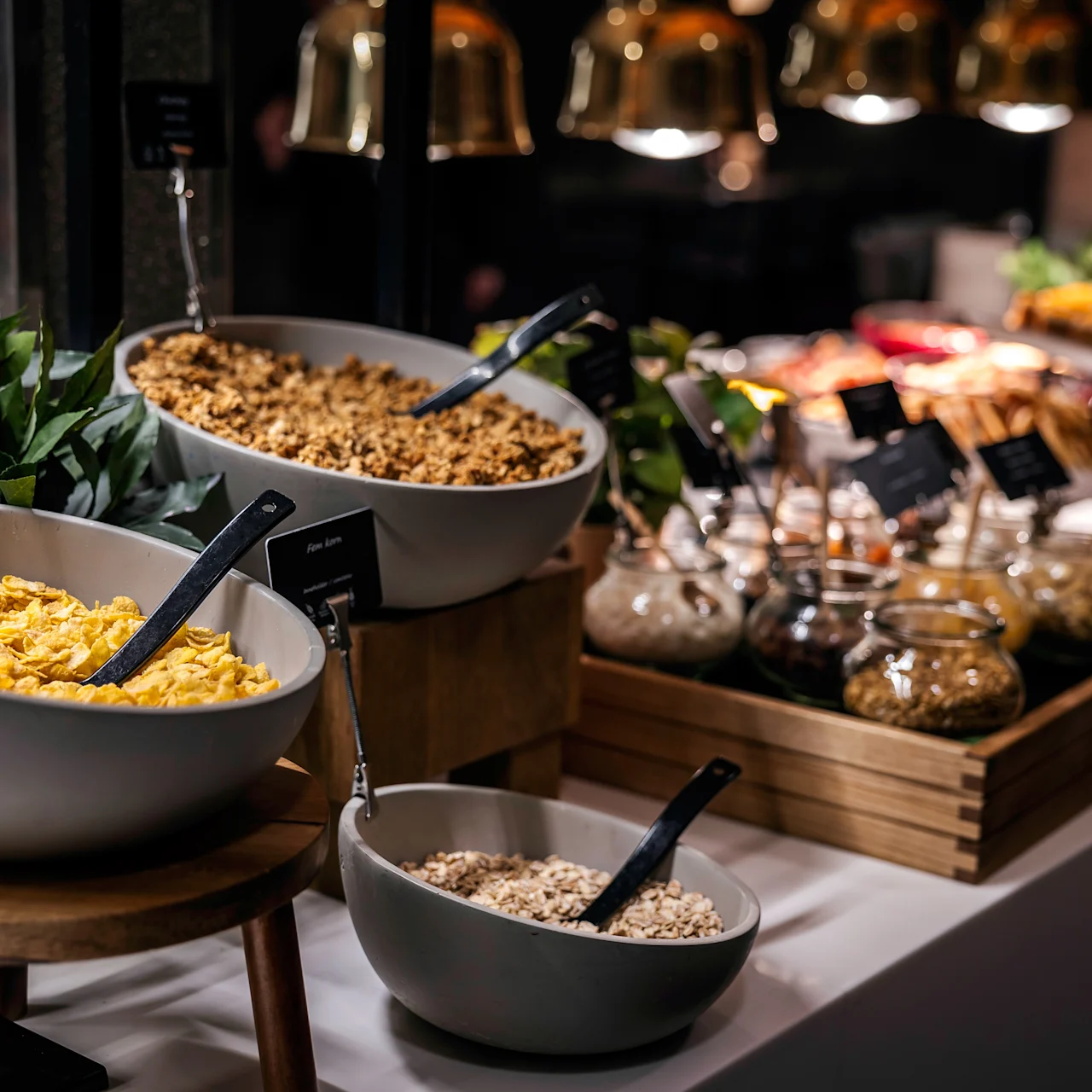 Bowls of cereals and serving spoons are displayed on a breakfast buffet, surrounded by jars of condiments and greenery. Text: “Corn flakes” and “Plain Barley” labels are visible.