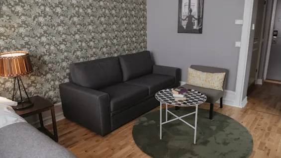 Gray sofa situated against floral-patterned wallpaper, complemented by a small side table holding a book, and an adjacent armchair. Wooden floor and a cozy rug complete the room's ambiance.