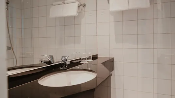 Ett rent och modernt hotellbadrum med mörk bänkskiva, vitt handfat, glänsande kranar och fräscha vita handdukar på en ställning.