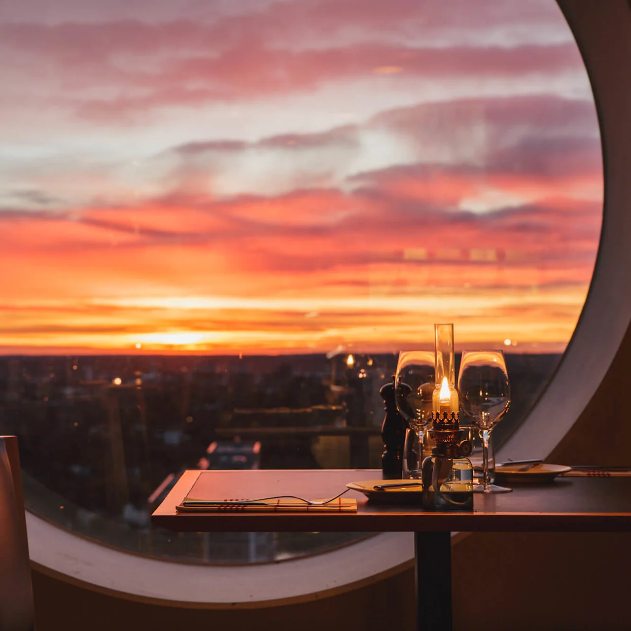 Et bord dækket til middag med et lys og glas står ved et stort rundt vindue, med en livlig solnedgang i orange og pink i horisonten.
