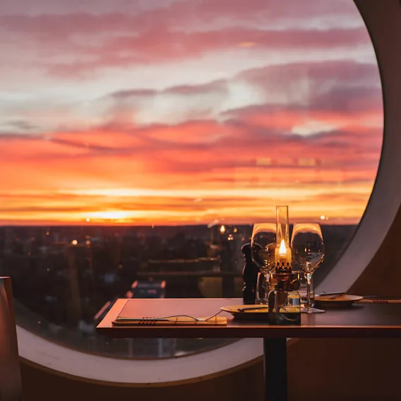 Ett bord dukat för middag med ett ljus och glas står nära ett stort runt fönster, med en livlig solnedgång som kastar nyanser av orange och rosa på himlen.