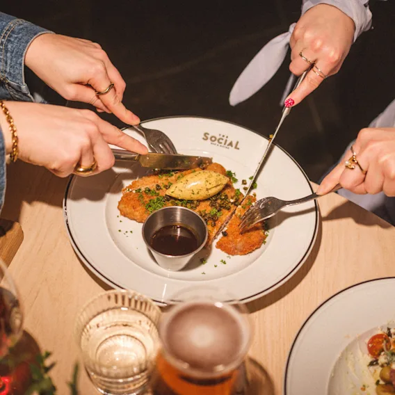 Hands cutting a shared meal on a plate with the word SOCIAL, enjoying a dining experience together.