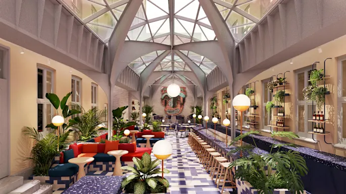 Dining area with colorful seating and tables; lush plants and modern lighting; surrounded by large windows and high vaulted ceiling; vibrant wall sculpture at the back; stylish, urban atmosphere.