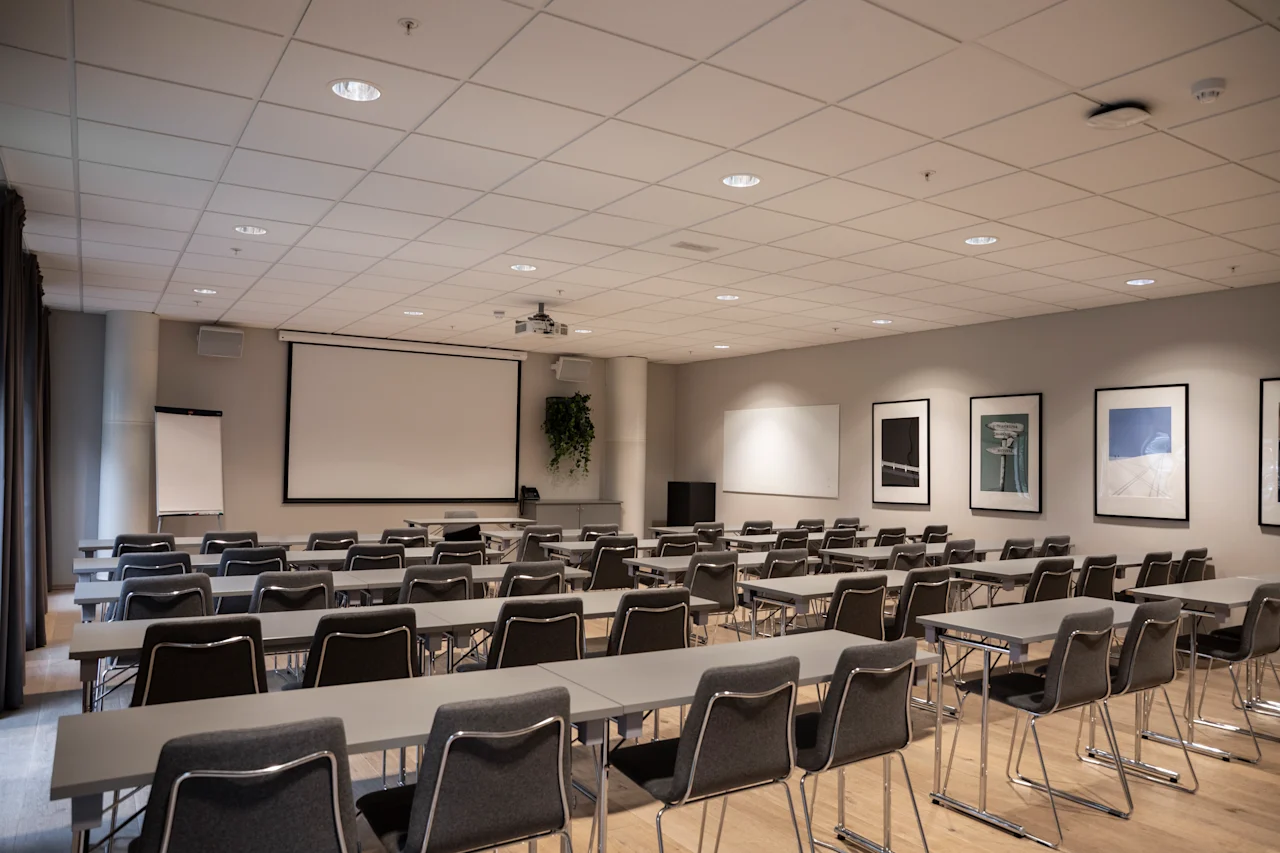 Et konferanserom er satt opp med rader av tomme stoler vendt mot en stor projeksjonsskjerm. Veggene er dekorert med innrammet kunst, noe som skaper et profesjonelt og organisert miljø.