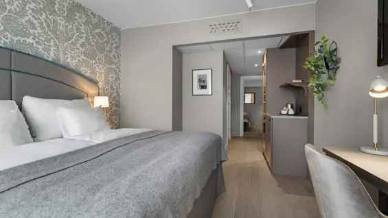A modern hotel room with a comfortable bed, patterned wall, desk, and a small kitchenette.