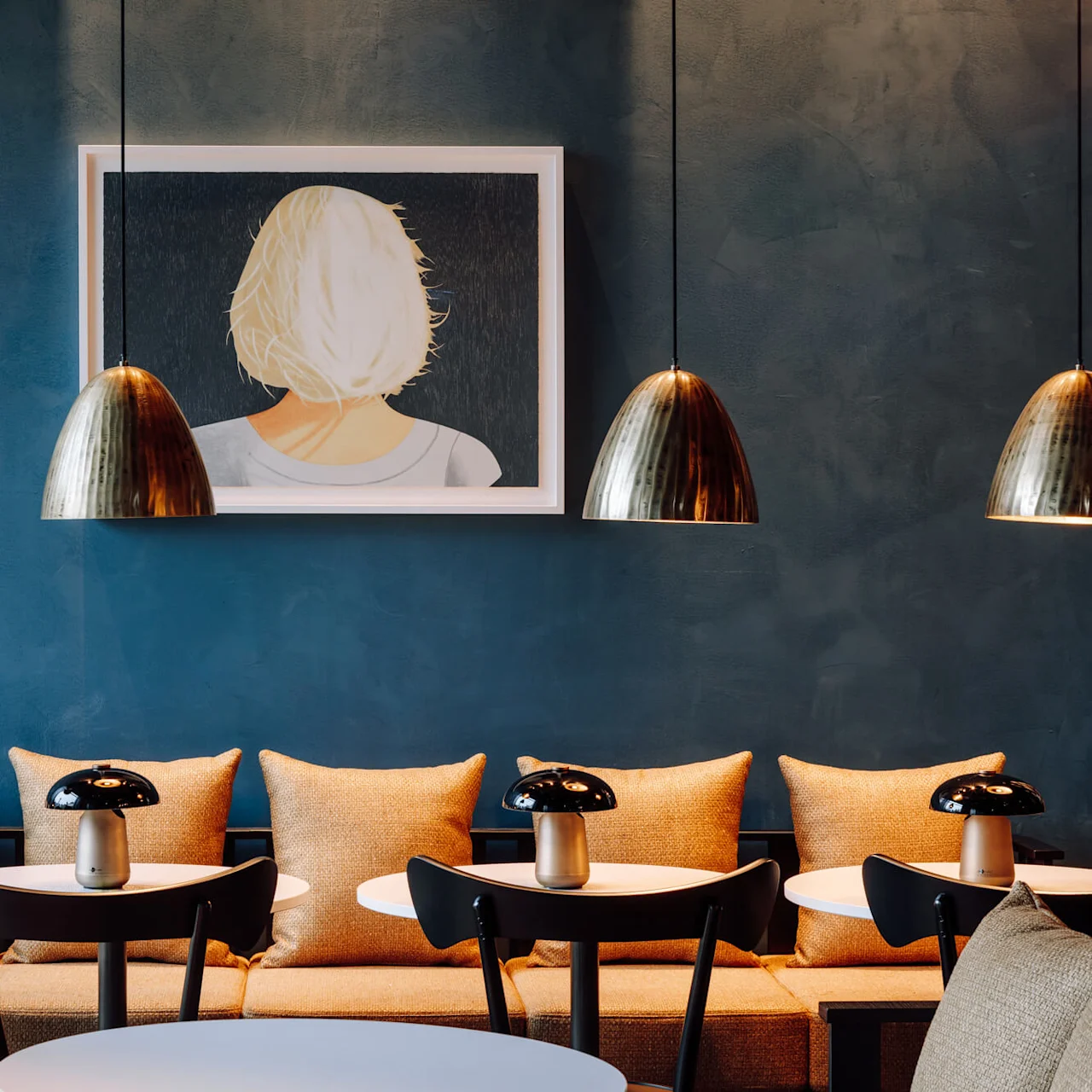 Artwork of a woman with blonde hair hangs on a teal wall. Three pendant lamps illuminate a cozy seating area with cushioned chairs below.