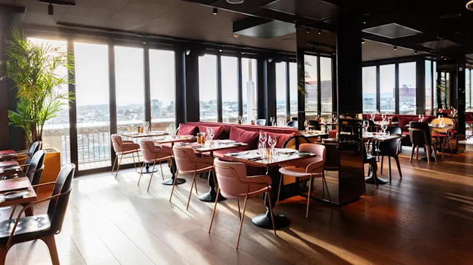 A bright, modern restaurant with wooden floors features pink and black chairs around set tables. Large windows provide a city view, and lush green plants enhance the elegant atmosphere.