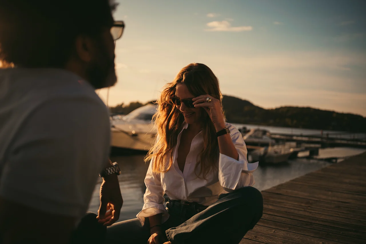 To personer nyder en samtale på en træmole ved solnedgang, med både i baggrunden.