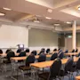 Et konferencelokale med rækker af borde og stole vendt mod en stor projektorskærm og et podium, klar til en præsentation.