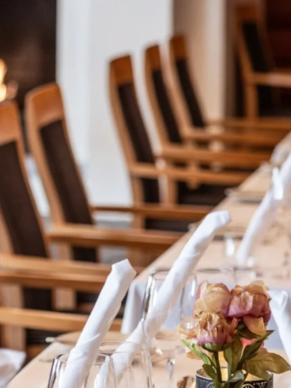 Et bord dekket med vinglass og rullede hvite servietter, med en liten vase med rosa blomster, plassert i en formell spisesal med trestoler.