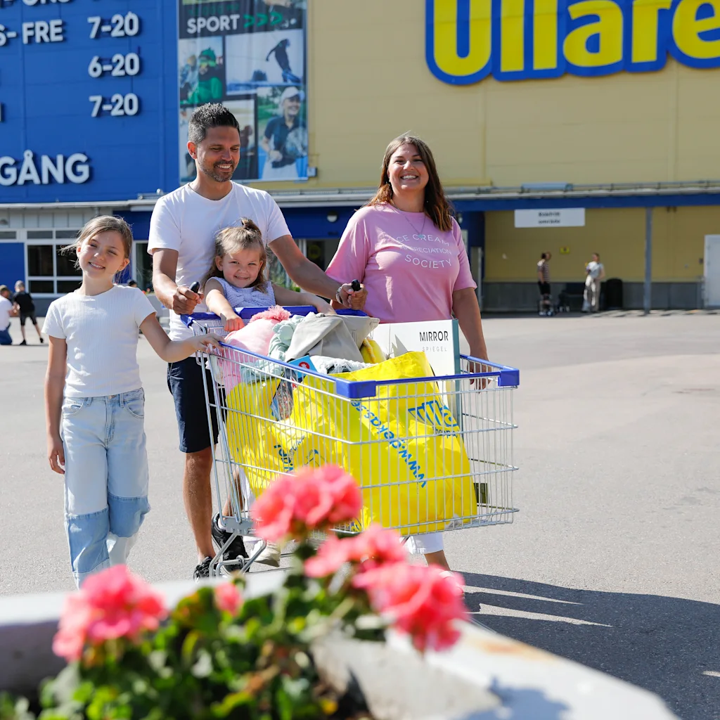 Family leaving Gekås Ullared with a full shopping cart. Store hours: TORS-FRE 7-20, LOR 6-20, SON 7-20. UTGANG.