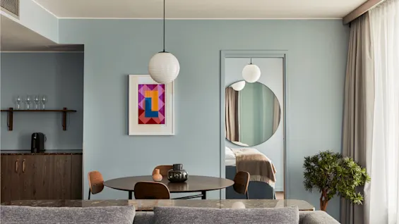 A round table with wooden chairs is situated in a modern living room. A circular mirror and artwork adorn the light blue wall, with a plant beside the window.