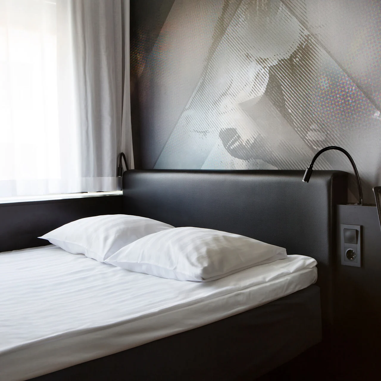 Et moderne hotelværelse med en pænt redt seng med hvide lagner og puder, placeret ved siden af et vindue med tynde gardiner og belyst af vægmonterede læselamper.