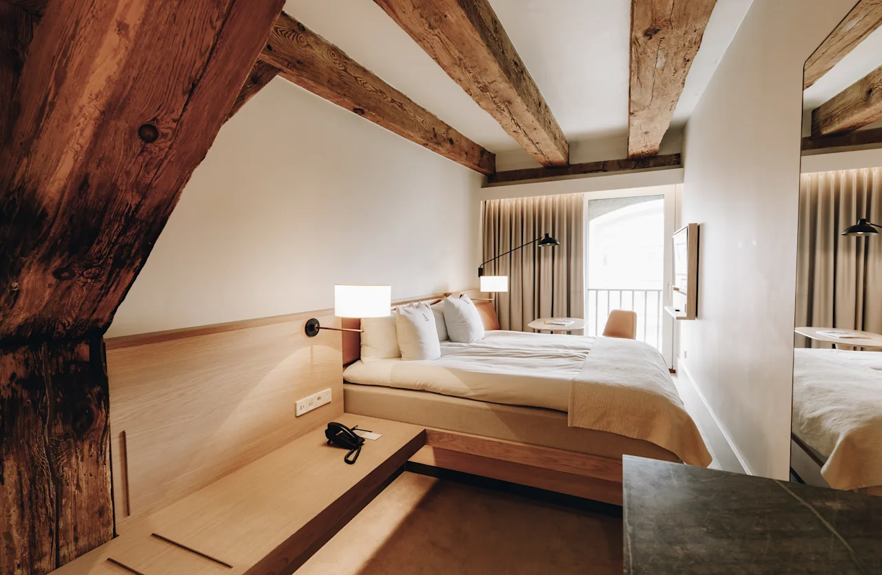 Ett mysigt hotellrum med en bekväm säng, synliga träbjälkar och en ljus balkongutsikt, för en avkopplande vistelse.
