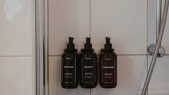 Tre mørke flasker med body wash, shampoo og balsam monteret på en flisebelagt brusevæg, klar til brug.