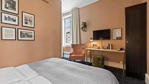 Et komfortabelt hotellrom med en seng, et skrivebordsområde med TV, og et vindu, dekorert med innrammede bilder på veggen.