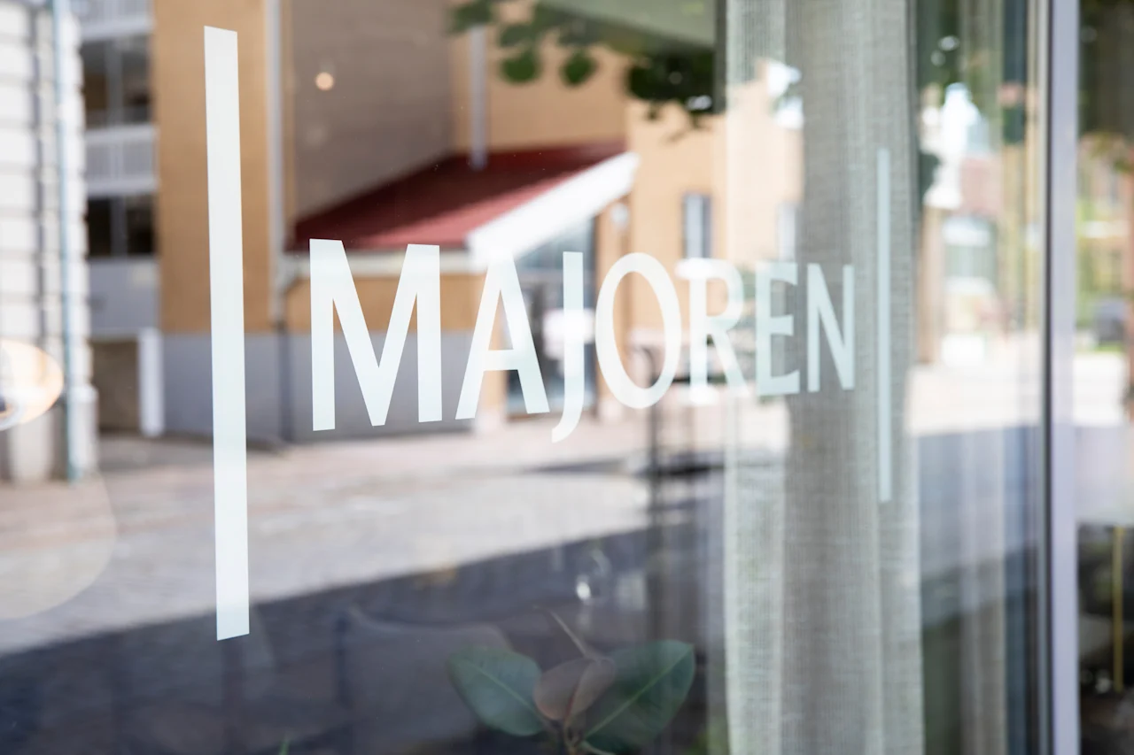 Lähikuva lasisisäänkäynnistä, jossa näkyy sana MAJOREN, heijastaen sumeaa katunäkymää ja rakennusta.