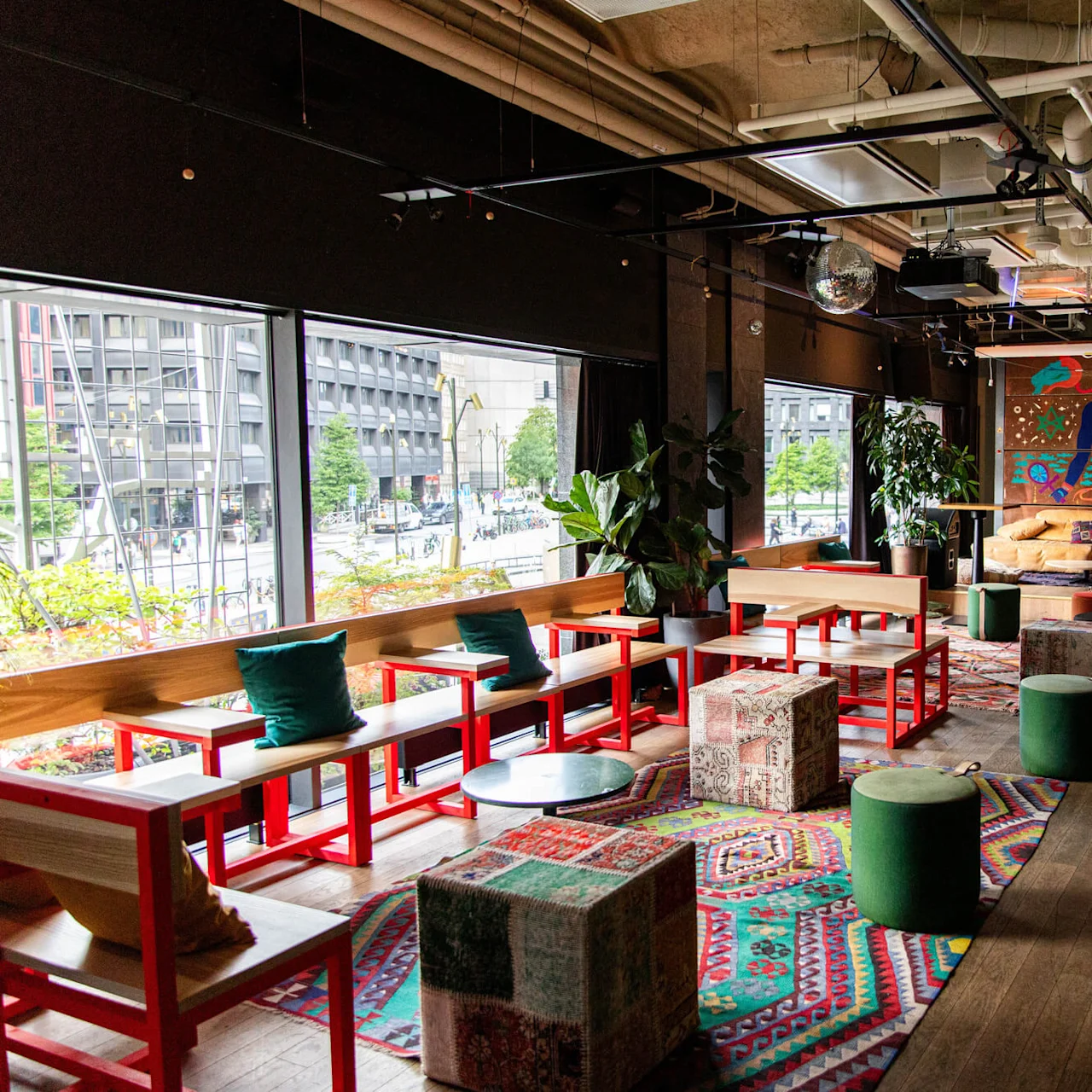 Colorful lounge area featuring vibrant furniture and abstract art. Bright red tables and green stools sit on patterned rugs, with large windows overlooking an urban street scene.