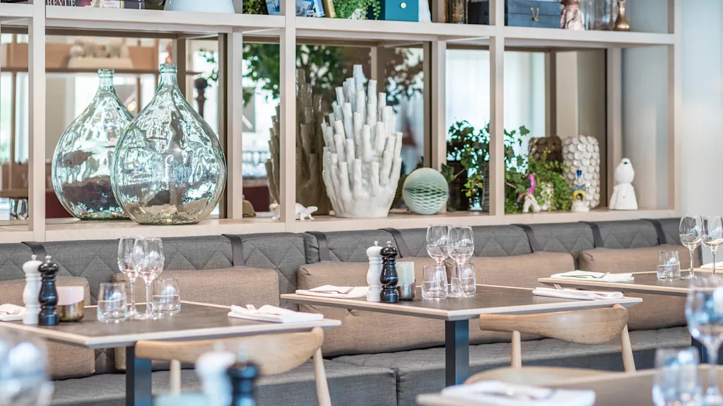 Restaurangbord är dukade med glas och servetter på gråa vadderade bänkar. Bakom pryder dekorativa glasflaskor och växter hyllor av trä och skapar en modern och luftig atmosfär.