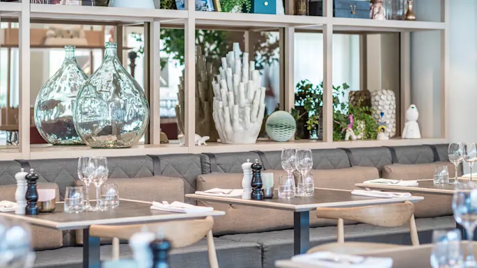 Restaurantbord er dækket med glas og servietter på grå polstrede bænke. Bagved pryder dekorative glasflasker og planter træhylder, hvilket skaber en moderne og luftig atmosfære.