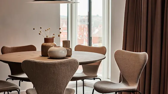 Pyöreä pöytä pitää koristeellista keramiikkaa ja kuivattuja kukkia tumman riippuvalaisimen alla, neljän beigen tuolin ympäröimänä. Lattiaan asti ulottuvat ikkunat ja ruskeat verhot kehystävät huonetta.