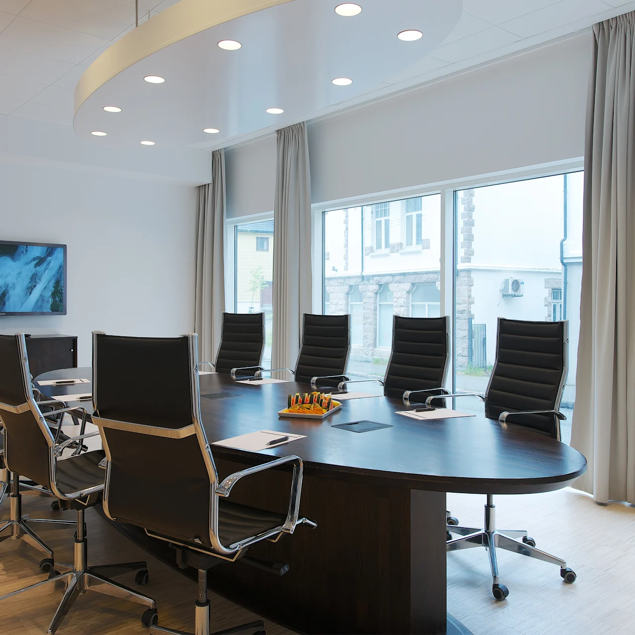Ett stort, oval konferensbord omges av svarta stolar i ett kontor med stora fönster. En väggmonterad skärm visar en bild av ett vattenfall, under ett tak med cirkulära lampor.