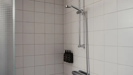 A clean and modern shower area with white tiled walls, a chrome shower fixture, and wall-mounted dispensers. Home Hotel Uppsala.