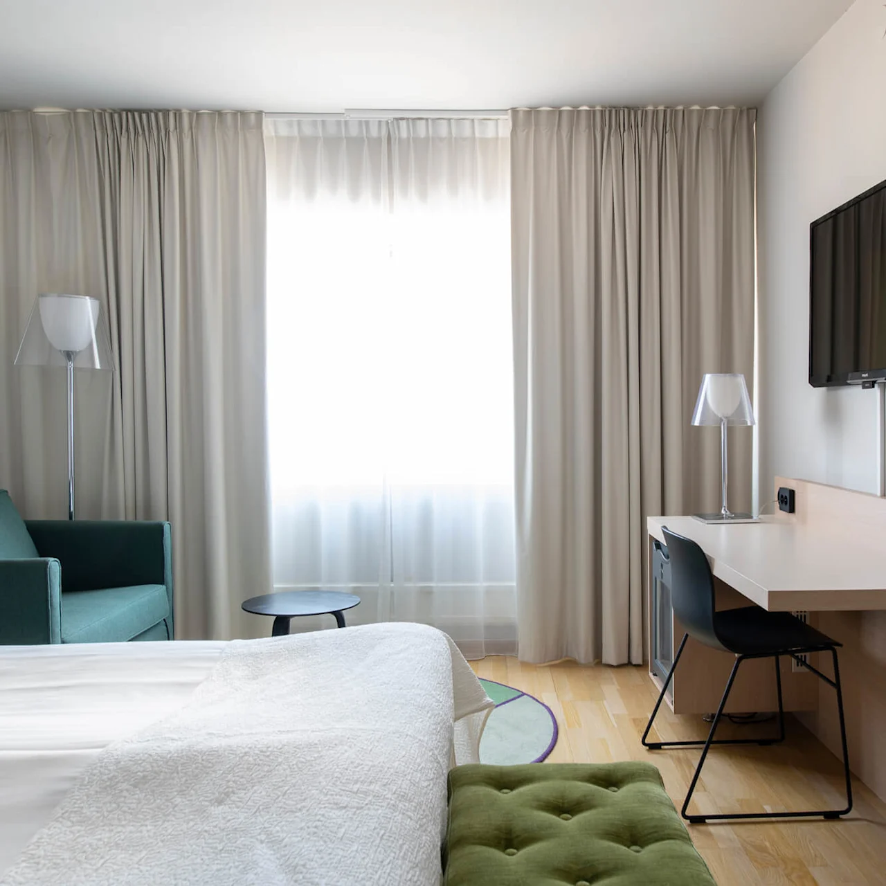 En säng med vita sängkläder står i ett hotellrum. Den vetter mot ett skrivbord med stol, en väggmonterad TV och ett fönster med beige gardiner. En grön soffa står i närheten.