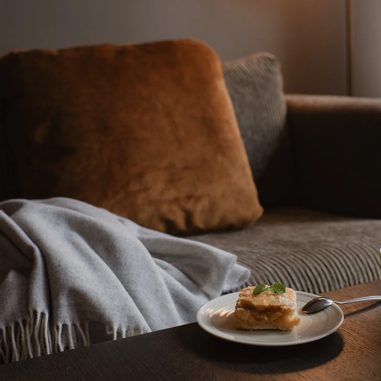 A plate with pastry rests on a table beside a steaming mug, in front of a cozy sofa draped with a blanket and a large cushion.