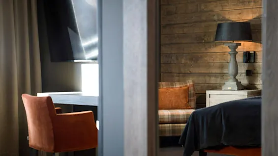 Et hyggeligt rum har en ternet sofa med en orange pude, ved siden af et natbord i træ med en lampe. Et skrivebord med en rustfarvet stol står under dæmpede gardiner.