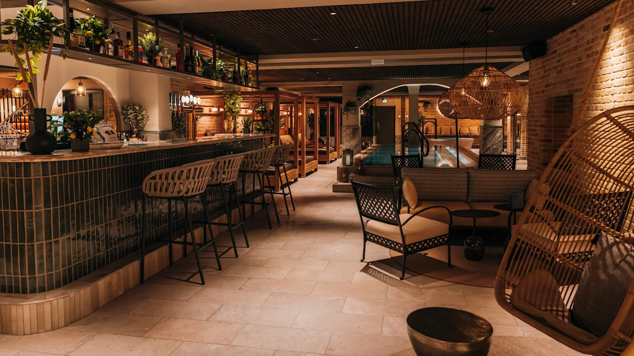 Bar with high stools, backed by shelves with plants and bottles; adjacent to a cozy seating area and illuminated indoor pool in a warmly-lit modern lounge setting. 