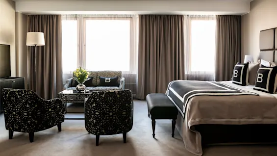Et stilfuldt hotelværelse har en seng med sort og hvidt sengetøj, et siddeområde med mønstrede stole og en sofa med "B"-puder. Store vinduer omkranser rummet.