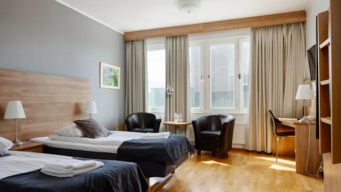 To pent oppredde enkeltsenger med lamper på sidebord, plassert i et hotellrom med tregulv og store vinduer. Stoler og et lite bord står ved vinduet.