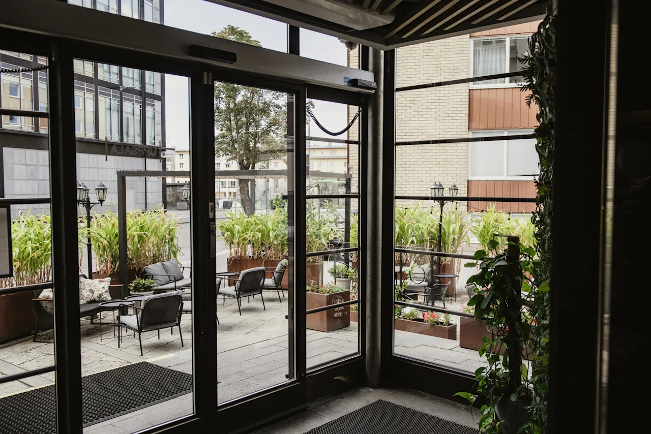 View from inside Home Hotel Uppsala looking out onto a cozy outdoor patio with seating areas and lush green plants.