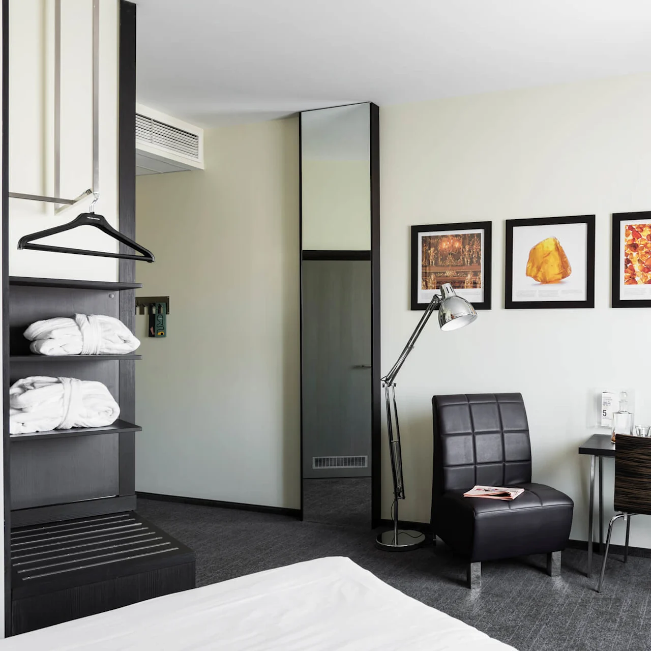 Ett hotellrum har en modern stol bredvid en lampa, nära en vägg med tre inramade konstverk. Ett skrivbord håller olika föremål, mittemot en liten tv på väggen.