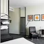 A hotel room features a modern chair beside a lamp, near a wall with three framed artworks. A desk holds various items, opposite a small television on the wall.