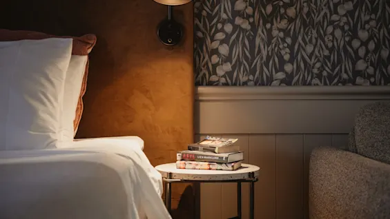 A comfortable reading nook featuring a bed, a side table with books including ASYL and VILSE, and soft lighting.